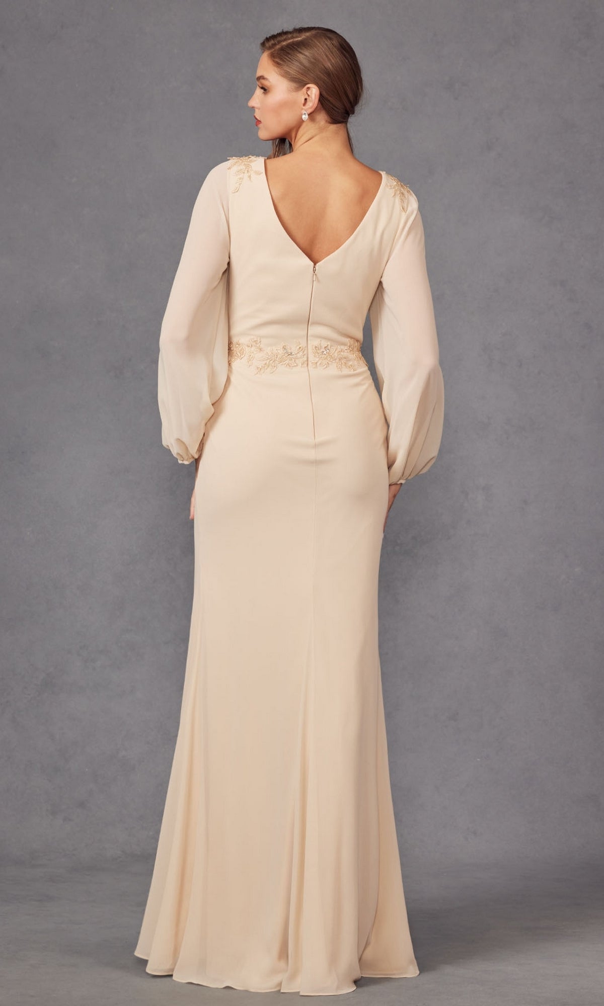A woman stands facing away in the Juliet JTM16A V-Neck Long Sleeve Prom Gown, showcasing its deep V-shaped back and light beige color against a plain gray background.